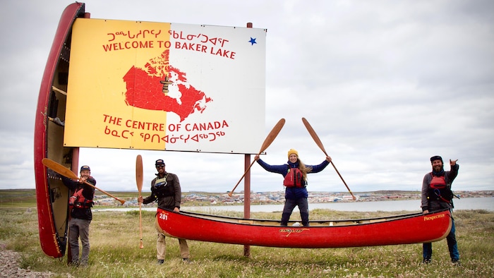 Les trois aventuriers et une femme posent avec leurs canots devant une affiche sur laquelle est inscrite « Bienvenue à Barker Lake, le centre du Canada »