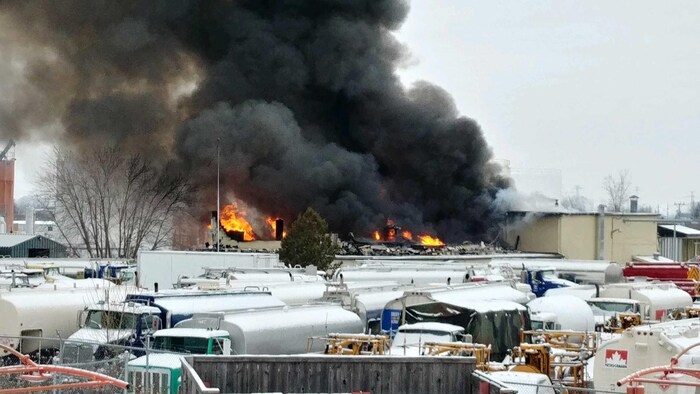 À l'arrière-plan de plusieurs camions-citernes garés près de bâtiments commerciaux s'élèvent des flammes et de fumée noire.