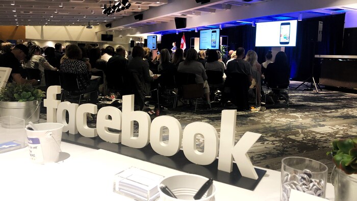 Vue sur une grande salle. Il y a environ 300 personnes. Elles écoutent une représentante de Facebook qui est sur une scène. À l'avant-plan, on voit le logo de Facebook.
