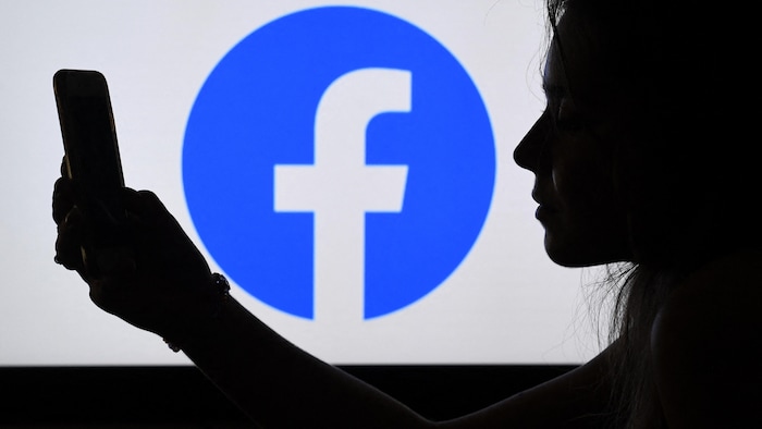 Une femme regarde un téléphone. Le logo de Facebook est derrière elle.