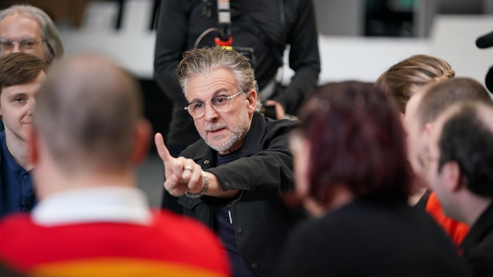 L'acteur lève un doigt en l'air en parlant à plusieurs personnes assises à ses côtés. 