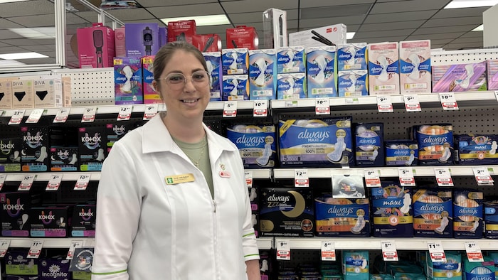 Une jeune femme en sarrau sourit à la caméra. Elle se trouve debout dans une allée de produits menstruels.