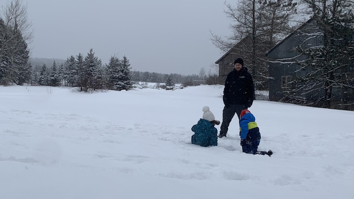 Un homme joue avec deux enfants, dans la neige d'un secteur boisé, à la campagne.