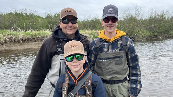 Un père et ses deux fils habillés pour la pêche. Ils posent pour une photo à côté d'une rivière.