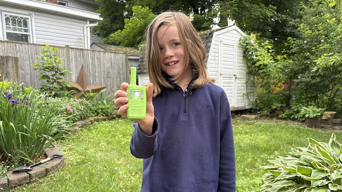 Un enfant debout dans la cour montre à la caméra un walkie-talkie vert fluo en plastique.