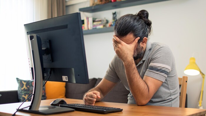 Un homme assis devant son ordinateur se touche le front car il a mal à la tête.