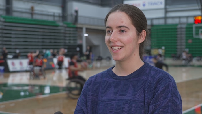 Une femme souriante en chandail bleu foncé s'exprime devant un terrain de basketball en fauteuil roulant.