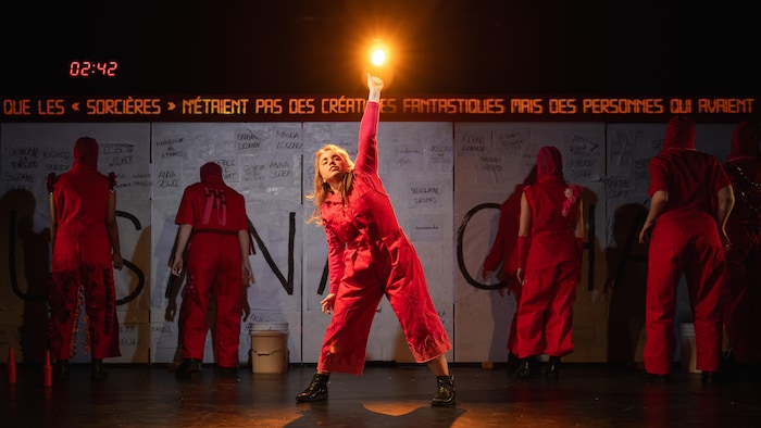 Une artiste en rouge pointe une lumière au centre d'une scène de théâtre entourée de performeurs devant un mur de noms.