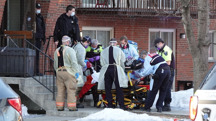 Une dizaine de membres des services d'urgence s'active à l'entrée d'un immeuble à logements. Une bonne partie d'entre eux transportent une personne sur une civière.