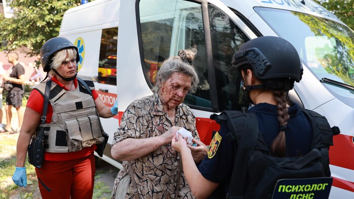 Debout à côté d'une ambulance, une femme blessée à la main, avec le visage ensanglanté, reçoit des soins de deux ambulancières qui portent des casques de protection et des gilets pare-balles.