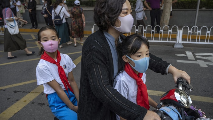 Une femme tient le guidon de sa mobylette. Une enfant est assise en avant d'elle, et une autre en arrière.