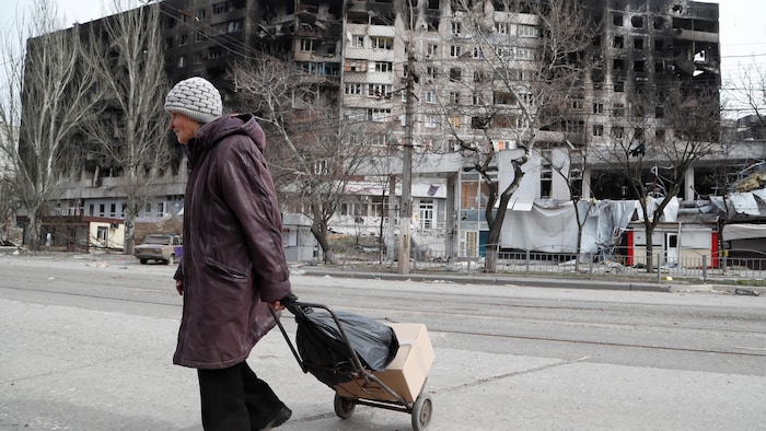 Une femme âgée marche avec un chariot devant un grand immeuble de logements endommagé par les bombes.