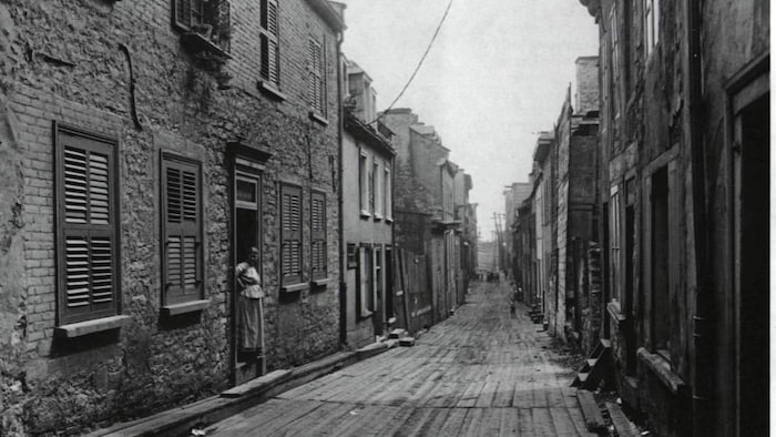 Une femme se tient dans l'embrasure de sa résidence, dans une rue en enfilade, dont la voie est en planches de bois.