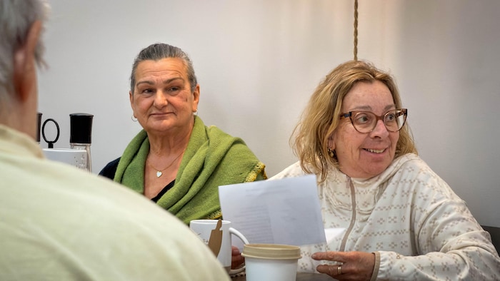Deux femmes écoutent d'autres membres de la chorale discuter, autour d'une table. 