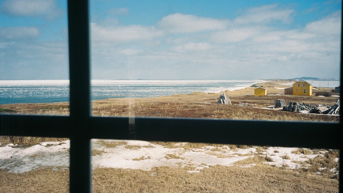Un plan très rapproché d'un carreau de fenêtre qui laisse voir une bande de terre bordée des deux côtés par la mer, à Pointe-aux-Loups, aux Îles-de-la-Madeleine.