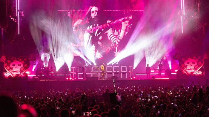 Vue d'ensemble d'un performance  d'Avril Lavigne aux Plaines d'Abraham, le 4 juillet 2025. Avril Lavigne se tient au centre de la scène, entourée de musiciens et de projections lumineuses roses et blanches. Un expansive  écran affiche des visuels en noir et blanc d'elle-même jouant de la guitare, et des motifs de crânes et d'os croisés ornent les côtés de la scène. Une vaste foule d'admirateurs, dont plusieurs filment avec leurs téléphones, remplit le premier plan.