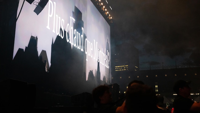 Un écran de concert géant affiche en blanc le texte "Plus chaud que Montréal?" sur un fond sombre de silhouettes de bâtiments. Des lumières de scène sont visibles au-dessus de l'écran, et les têtes sombres du public sont visibles en bas de l'image, avec un ciel nuageux ou enfumé en arrière-plan.