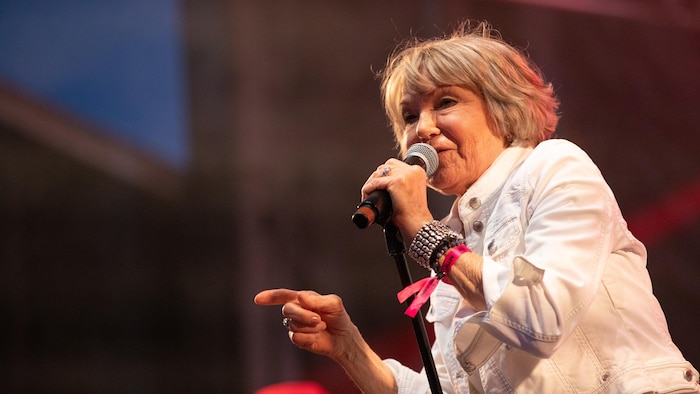 Plan rapproché de la chanteuse québécoise Marjo sur scène, le 4 juillet 2025. Elle tient un microphone de la main droite et pointe l'index de sa main gauche vers la gauche. Ses cheveux sont courts et blonds, et elle porte une veste blanche en jean par-dessus un haut sombre.