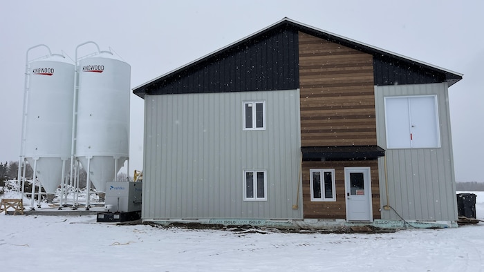 La photo présente un bâtiment de ferme moderne sous la neige, flanqué de deux silos blancs marqués "KINGWOOD". La façade combine revêtement métallique gris et bois, avec une porte blanche et quelques fenêtres sous un ciel couvert.