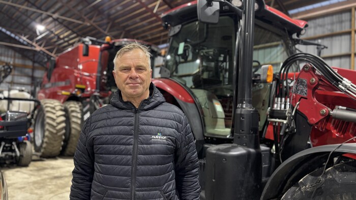 Un homme d'âge mûr portant une veste noire arborant le logo « Nutrinor Coopérative » pose devant d'imposants tracteurs rouges à l'intérieur d'un grand hangar agricole. Son expression est sereine et professionnelle.