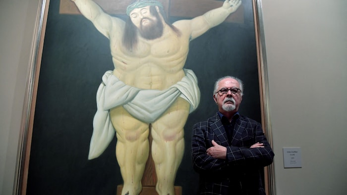 Le peintre se tient devant une peinture de Jésus sur la croix, dans le style de Botero avec des formes voluptueuses.