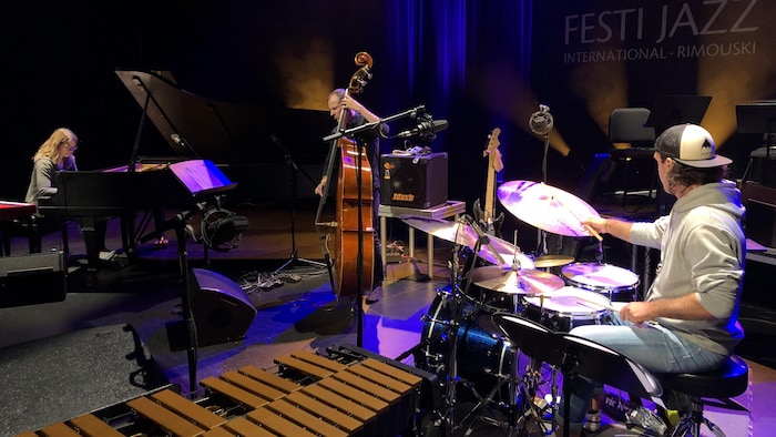 Emie R Roussel au piano avec un violoncelliste et un percussionniste derrière une batterie.