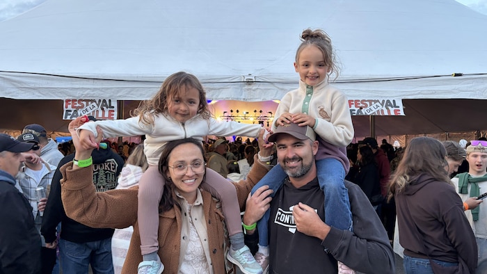 Un père et une mère portent fièrement leurs deux fillettes sur leurs épaules, tout sourire devant la grande tente ornée de la bannière du festival.