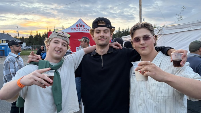 Trois jeunes hommes complices posent bras dessus, bras dessous, avec les tentes du festival et un début de coucher de soleil à l'arrière-plan.