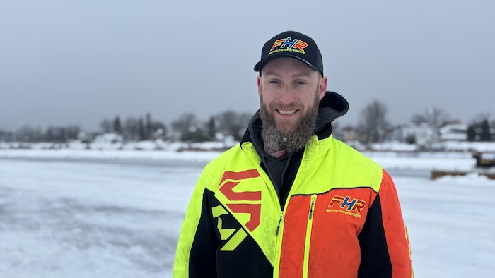 Un homme barbu sourit devant un paysage enneigé. Il porte une casquette et un manteau de motoneige aux couleurs vives (jaune, orange, noir) arborant le logo « FHR ». À l'arrière-plan, on aperçoit une étendue glacée et des habitations.