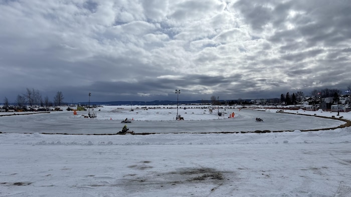 Cette photographie montre une course de motoneiges sur une piste ovale glacée en plein air. Le circuit est délimité par des bottes de foin sous un ciel lourd et couvert. À l'arrière-plan, on aperçoit des habitations et des arbres dénudés.