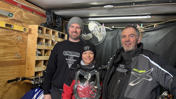 Trois personnes, deux adultes et un enfant muni d'une armoire de protection Fox, posent dans un garage ou une remorque. On aperçoit une moto bleue, des outils de menuiserie, un ventilateur et des vêtements de marque Ski-Doo en arrière-plan.