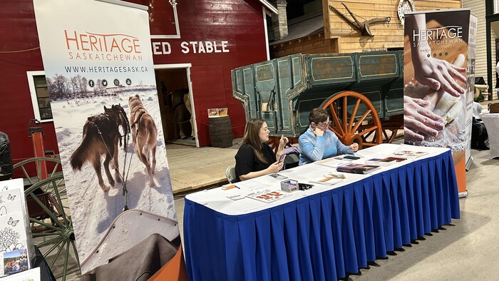 Deux personnes sont assises devant une table couverte de documents, avec, derrière elles, des pancartes sur lesquelles on peut lire le mot « Heritage », le 8 février 2026 à Saskatoon.
