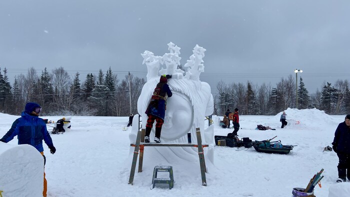 Des participants sculptent des formes diverses dans des blocs de neige. Une participante est juchée sur un échafaudage pour travailler sur le sommet d'une tête géante en neige.