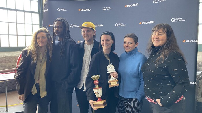 Six personnes posent devant un mur de commanditaires affichant les logos de Hydro-Québec et REGARD. Deux femmes au centre tiennent des trophées. Le groupe, vêtu de vêtements décontractés et variés, se tient debout dans un espace intérieur lumineux.