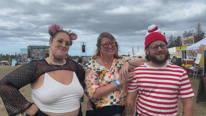 Sur le site d'un festival extérieur, deux femmes et un homme posent costumés. Le type est déguisé en Charlie avec un chandail rayé rouge et blanc et une tuque du même genre.