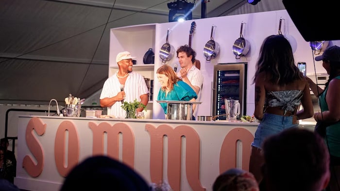 Les trois personnalités derrière un petit comptoir de cuisine aménagé sous une grande tente.