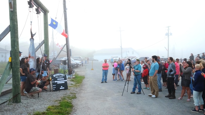 Des gens posent pour une photo sous un gros thon suspendu par la queue. Face à eux, des dizaines de personnes s'attroupent.