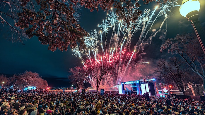 Un feu d'artifice jaillit derrière une scène où il y a une une immense foule.
