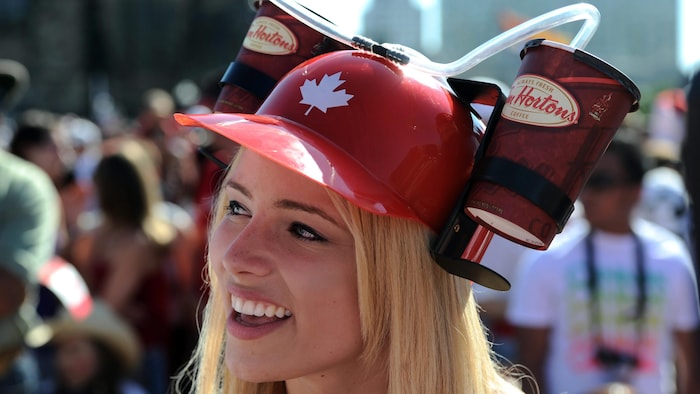 Femme coiffée d'un casque rouge en plastique orné d'une feuille d'érable, sur lequel sont attachés deux verres de Tim Hortons avec des pailles pour boire sans enlever le casque.