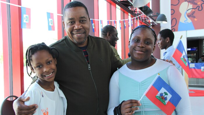 Un homme tient par les épaules ses deux filles. Les trois brandissent des drapeaux haïtiens en souriant à l'intérieur du Théâtre Cercle Molière à Winnipeg.