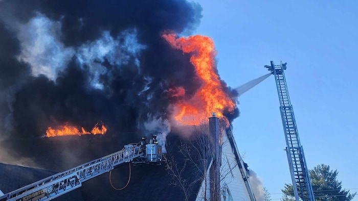 Des pompiers grimpés dans une échelle tentent d'éteindre le feu sur le toit.