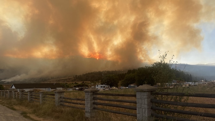 Un feu de forêt brûle sur une colline, projetant de hautes flammes et un immense panache de fumée orange et grise au-dessus d’une communauté. Au premier plan, on aperçoit une clôture, des habitations, des véhicules récréatifs et le capot d’une voiture.