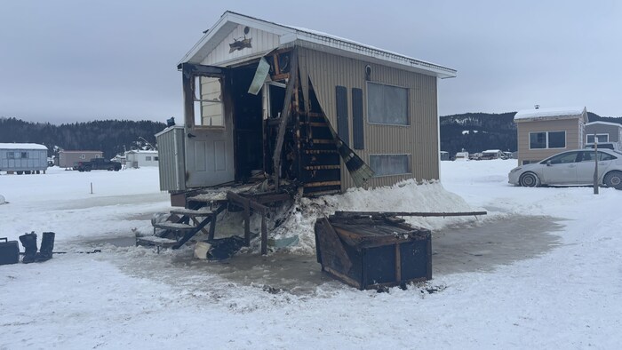 Cette photographie montre une petite cabane de pêche sur glace sévèrement endommagée par un incendie. La façade avant est calcinée et ouverte, révélant une structure brûlée. L'ensemble repose sur une surface enneigée et glacée sous un ciel couvert.