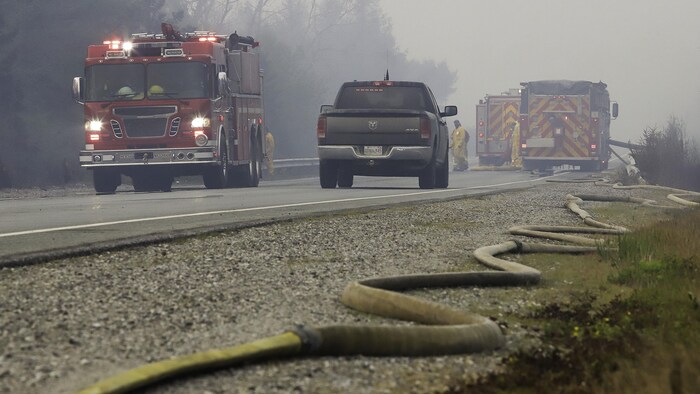 Un long boyau d'arrosage est placé en zigzag dans le gravier le long d'une autoroute. Des camions de pompiers, des camionnettes et des pompiers sont sur la route. La scène est voilée par de la fumée qui se dégage des feux de forêt.