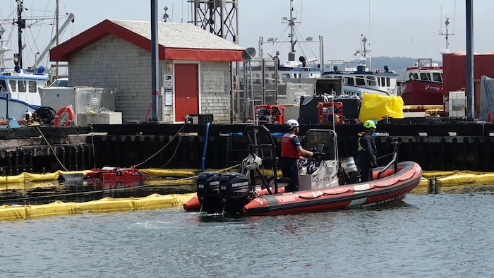 Des employés sur un Zodiac dans un havre de pêche près d'estacades flottantes devant un quai endommagé par le feu.