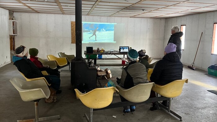 Des personnes sont assises et font face à une vidéo d'un skieur de fond à l'intérieur du chalet.