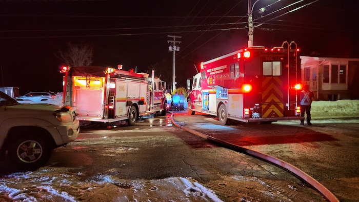 Deux camions de pompiers sont stationnés dans une rue, le soir. On voit un boyau d'arrosage sur le sol.