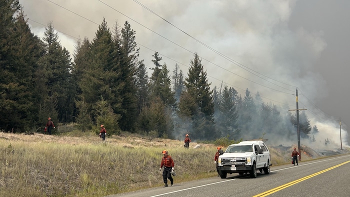 Des pompiers forestiers interviennent le long d’une route alors que de la fumée s’élève d’un secteur boisé touché par le feu de Pear Lake.