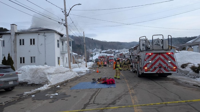 Cette photographie montre des pompiers en intervention devant un bâtiment résidentiel d'où s'échappe de la fumée. Plusieurs camions d'urgence bloquent la rue enneigée, délimitée par un ruban de sécurité jaune. Du matériel de secours est disposé sur une bâche bleue.