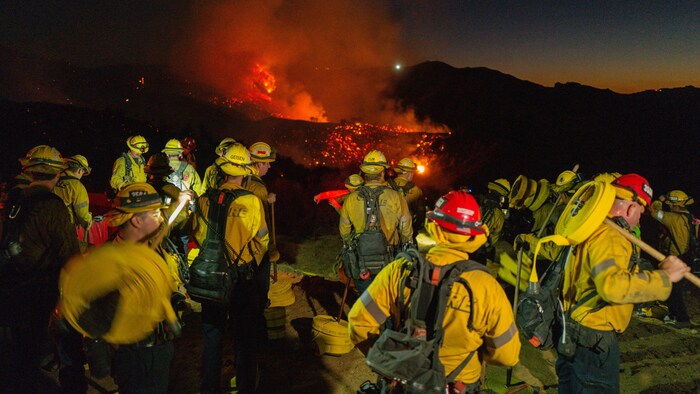 Des pompiers vêtus de jaunes avec des casques rouges devant un terrain en feu.